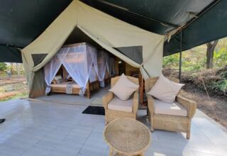 Luxury safari tent with tiled veranda, two cushioned wicker chairs and table, wooden beds draped in white mosquito nets.