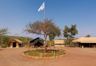 Canvas safari tents around a central tree and flag, with outdoor seating and dining in a rustic bushland camp.