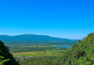Hillside view over a lush green valley to a shimmering lake and flat-topped mountain, small village below under clear blue sky