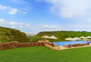 Infinity pool with sun loungers and umbrellas on a lawned terrace, overlooking a green valley and distant mountains.