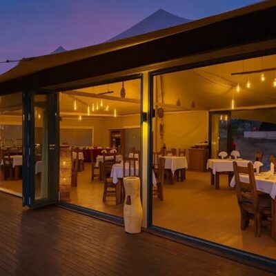 Open-sided tented dining room with wooden tables and white linens, warm pendant lights glowing onto a twilight deck.