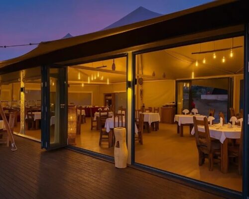Warmly lit open-air restaurant with wooden tables, white linens and pendant lights on a deck at twilight.