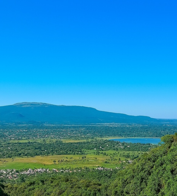 Lake and valley framed by lush hills, village rooftops below and distant mountain ridge under clear blue sky