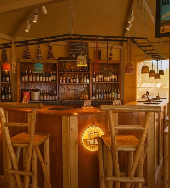 Rustic wooden hotel bar with high log stools, back shelves of bottles, woven pendant lamps and warm safari‑lodge vibe