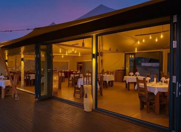 Warmly lit tented restaurant with open glass doors to a wooden deck, white-clothed tables and rustic wooden chairs set for dinner.