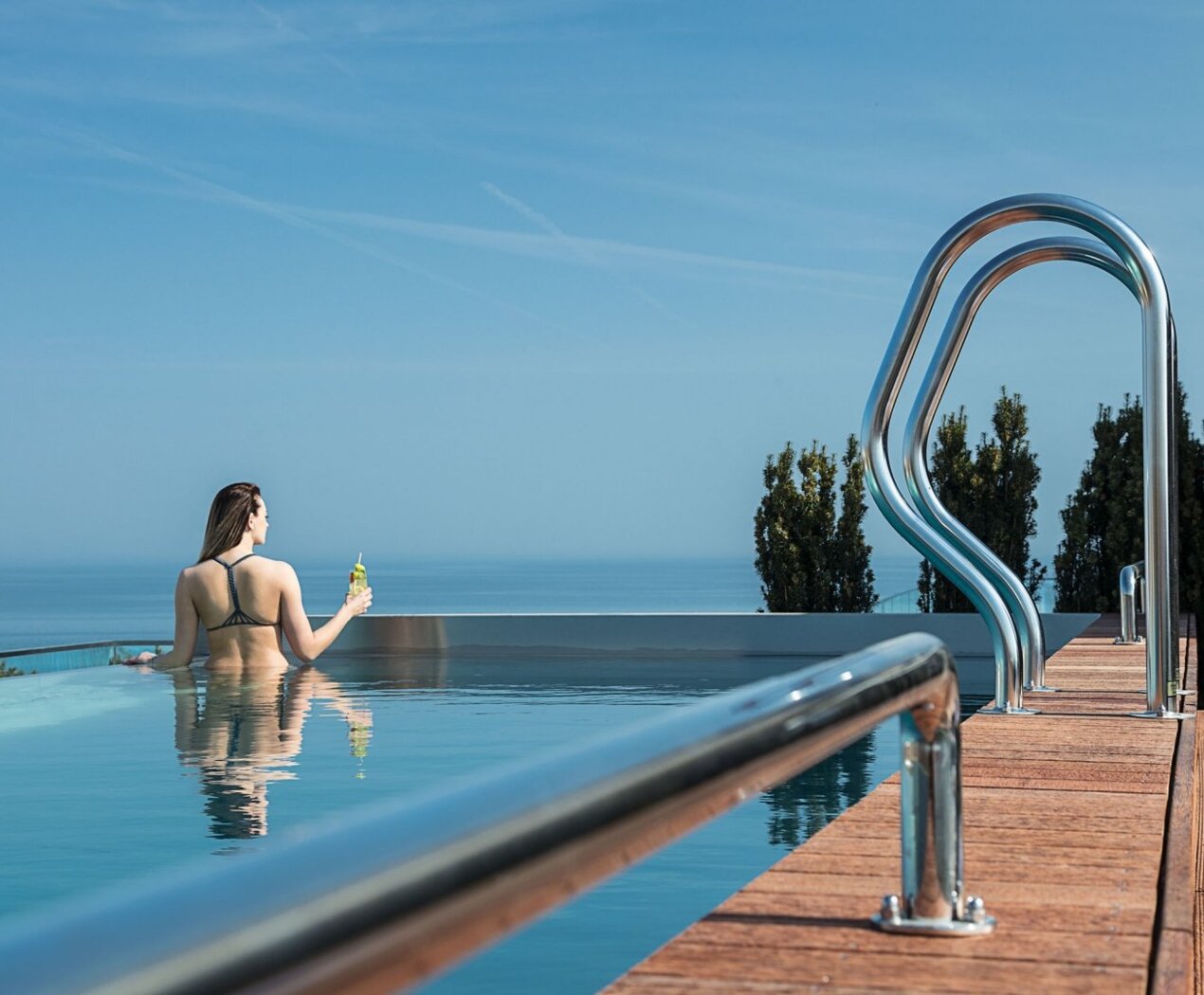 Guest relaxing in rooftop infinity pool with uninterrupted sea view, wooden deck and stainless handrails, holding a cold drink.