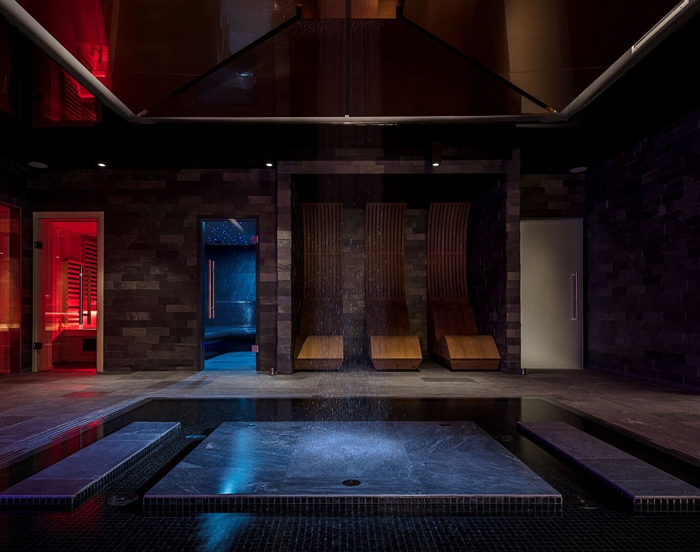 Luxurious hotel spa with central rain shower over stone platform, heated plunge pool, red-lit sauna and wooden lounge chairs.