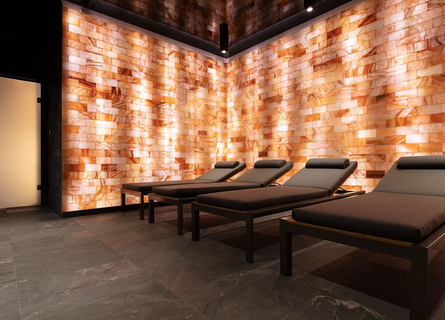 Spa relaxation room with four cushioned loungers, warm backlit Himalayan salt brick wall, slate floor and frosted glass door