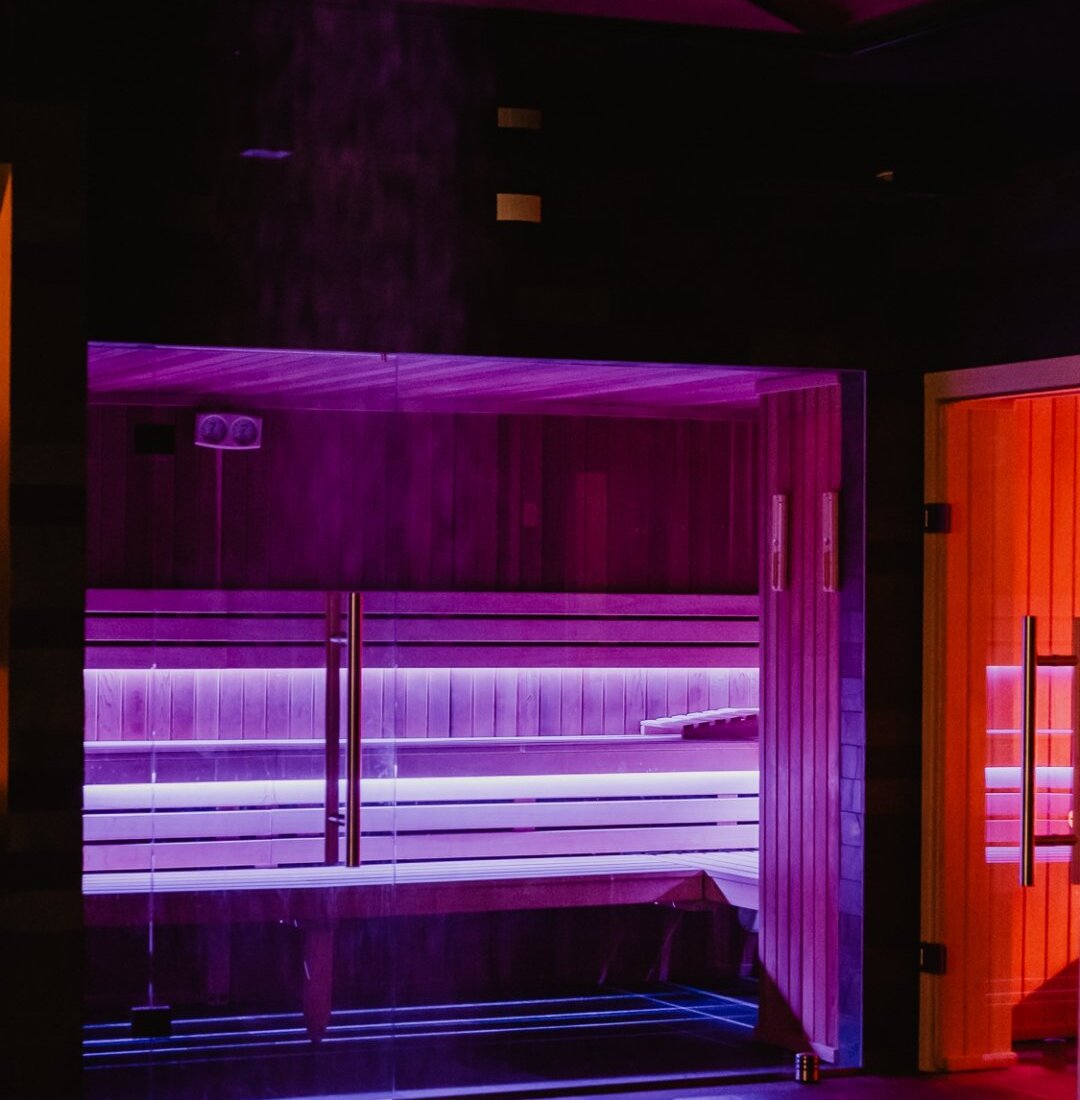 Glass-front sauna with wooden benches, purple LED mood lighting, adjacent orange-lit steam cabin and pool edge