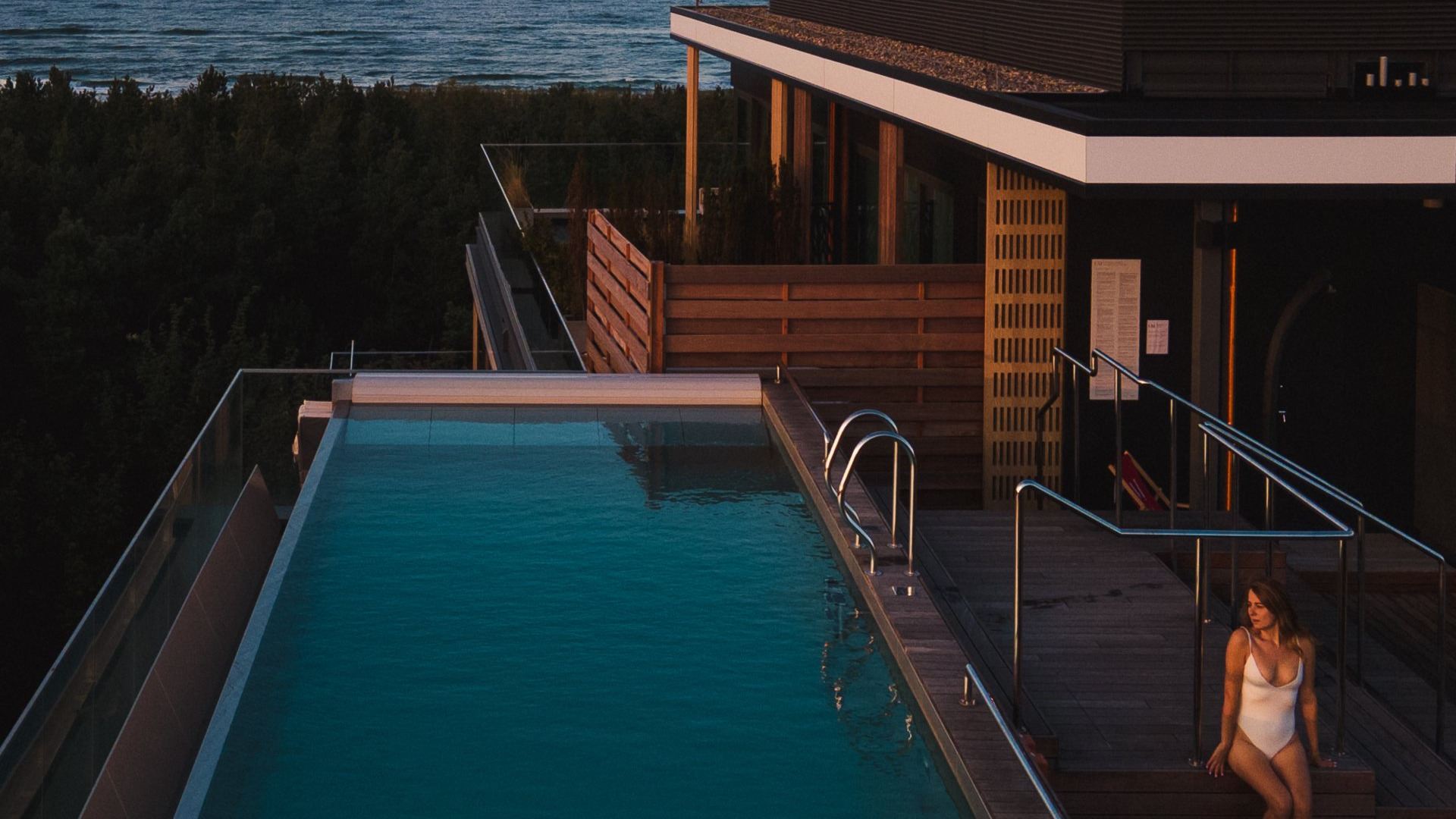 Rooftop infinity pool at dusk with ocean view, wooden deck and glass railing; guest in a white swimsuit seated on the steps.