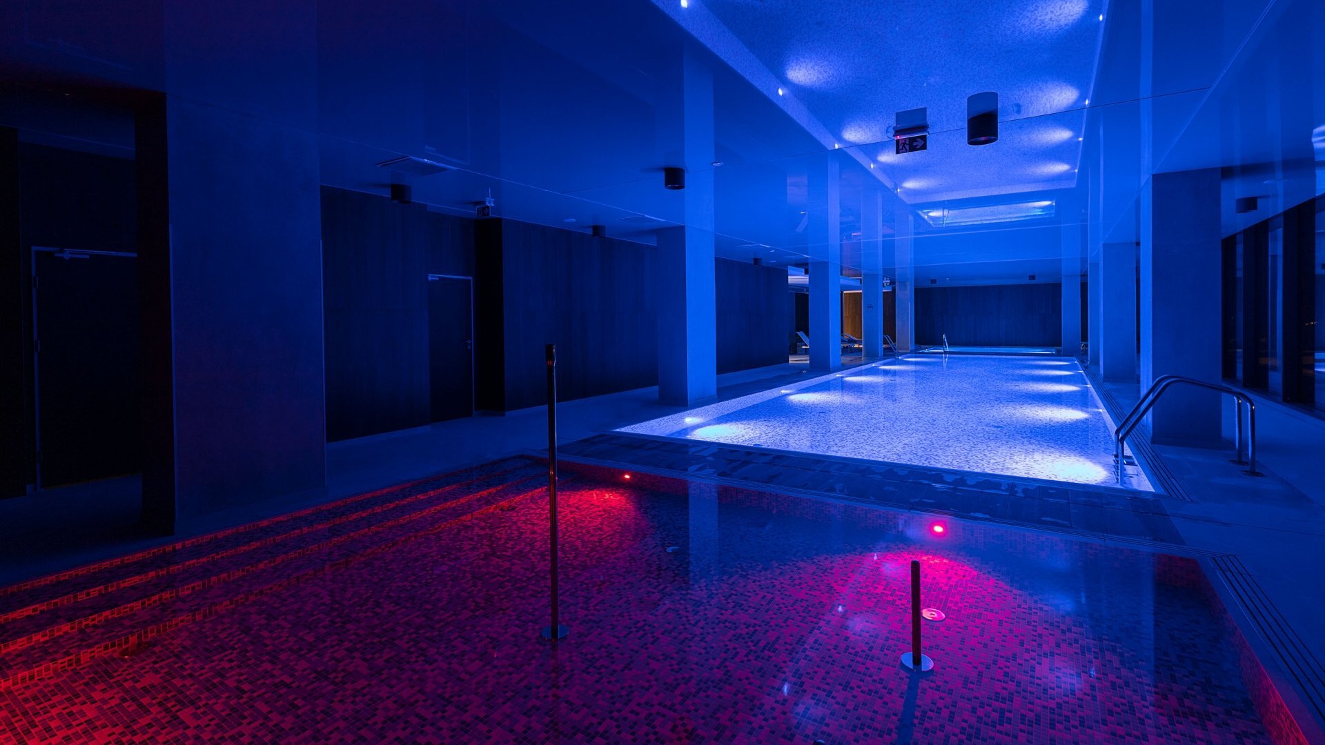 Indoor hotel pool with blue ambient lighting, mosaic spa lit red in foreground and large lap pool with stainless handrails