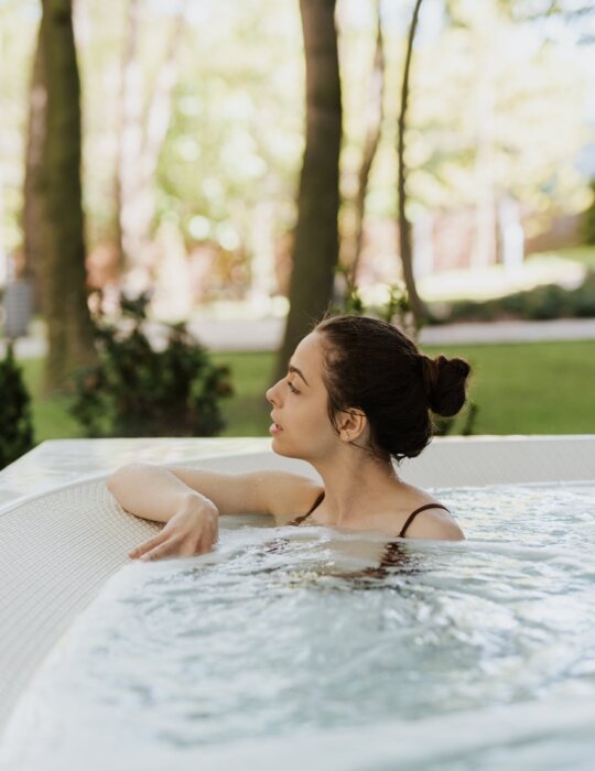 Woman relaxing in an outdoor hotel jacuzzi, leaning on the rim, warm bubbling water and garden trees visible for a peaceful spa atmosphere