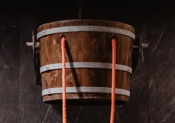 Wooden cold-shower bucket with orange pull rope mounted on a dark stone spa wall
