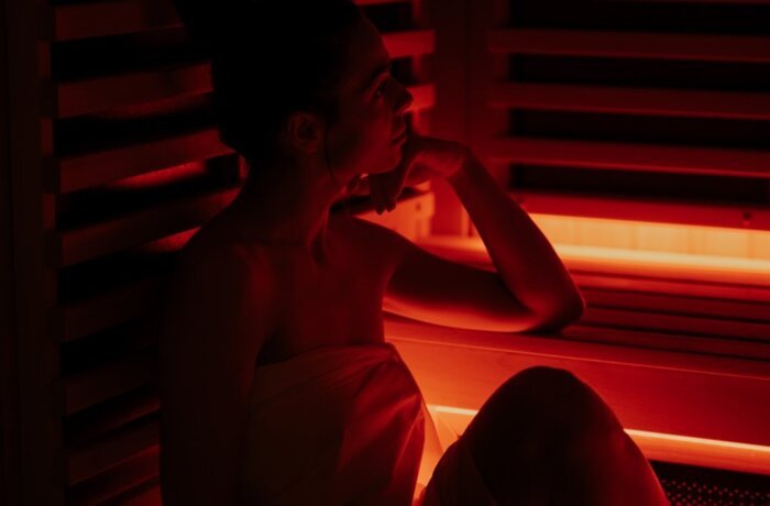 Frau im Handtuch entspannt in holzverkleideter Infrarotsauna bei warmem rotem Licht.