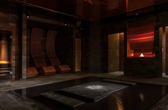 Dimly lit luxury spa with central heated plunge pool, falling water feature, wooden recliners, sauna room and glowing fireplace niche.