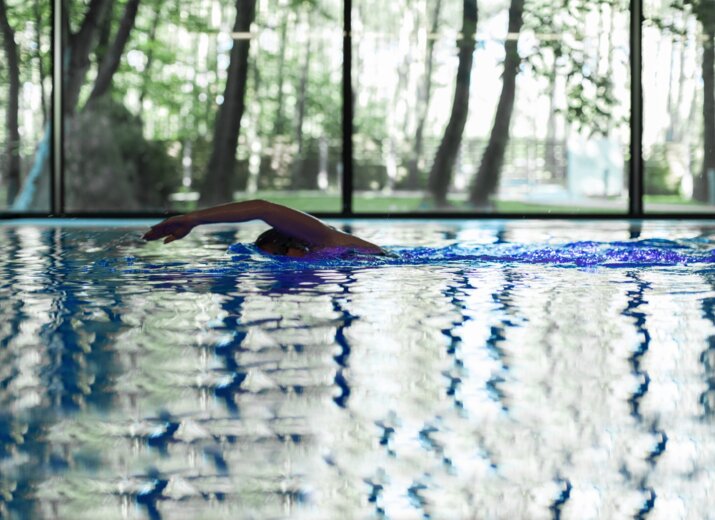 Swimmer doing laps in an indoor hotel lap pool with floor-to-ceiling windows and a peaceful forest view