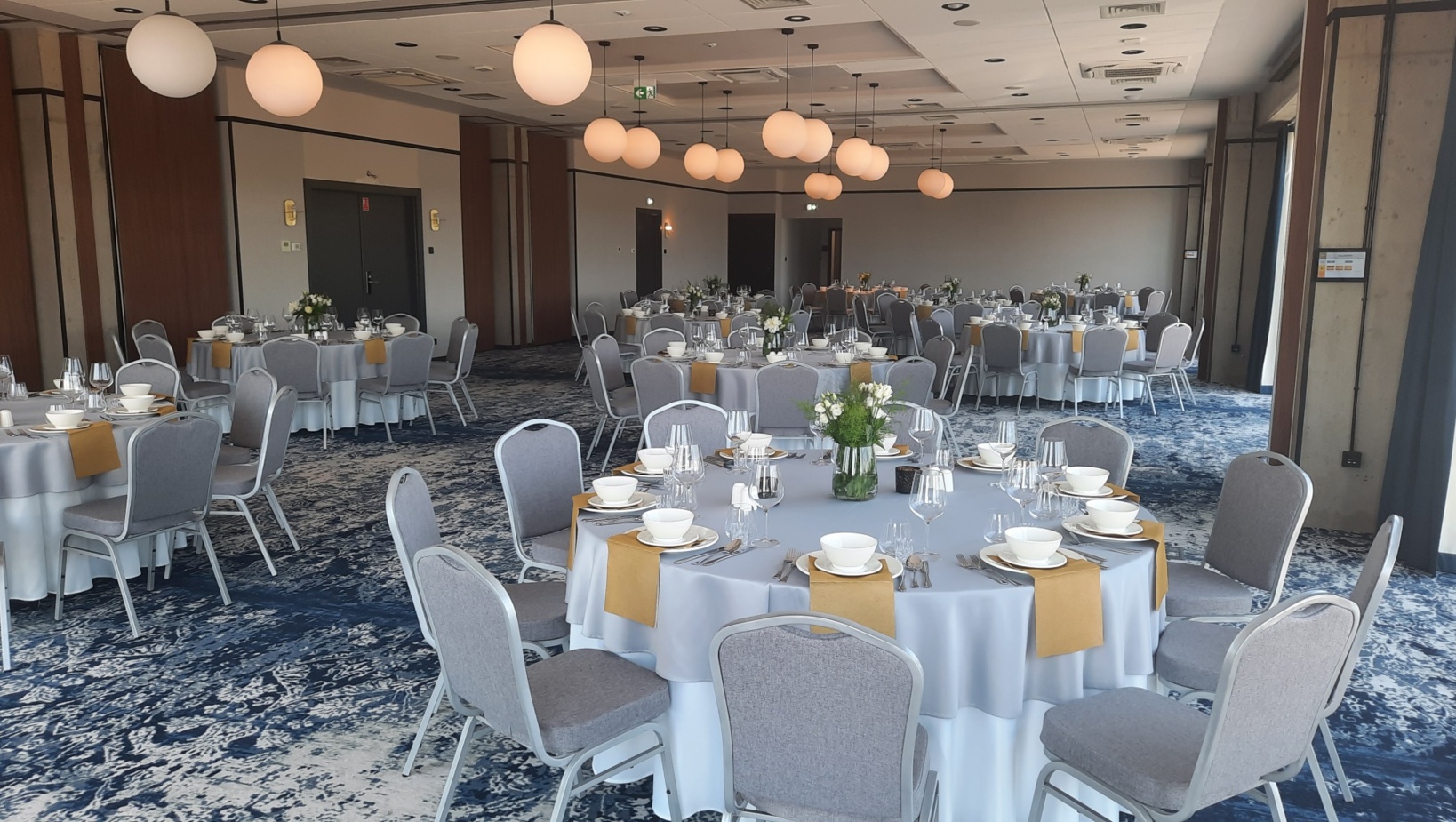 Elegancka sala bankietowa z okrągłymi stołami, szarymi krzesłami i dekoracjami kwiatowymi, przygotowana do uroczystej kolacji.