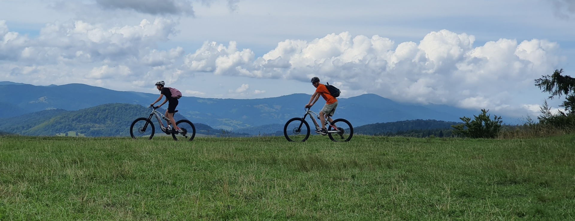 Dvaja cyklisti v prilbách a s batohmi idú cez zelený kopec, v pozadí horské pásmo a výrazné oblaky