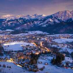 Zaśnieżona górska wioska z oświetlonymi, drewnianymi chatami o zmierzchu i panoramicznym widokiem na ośnieżone szczyty