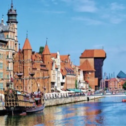 Historyczny nadbrzeżny deptak z ceglanymi kamienicami, kawiarnianymi tarasami, gośćmi spacerującymi i galleonem wycieczkowym.