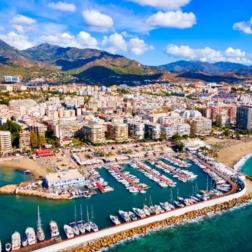 Coastal resort with marina and rows of yachts, sandy beaches, beachfront hotels and a mountain backdrop under blue sky