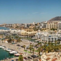 Marina with yachts, waterfront hotels, palm-lined promenade and blue sea with mountain backdrop.