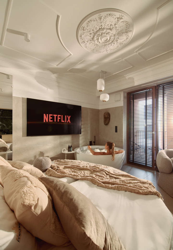 Luksusowy apartament z ozdobnym sufitem, przytulnym łóżkiem i wolnostojącą wanną; gość relaksuje się, oglądając Netflix.