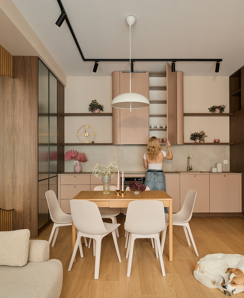 Aneks kuchenny w apartamencie z brzoskwiniowymi szafkami, drewnianymi panelami, stołem na sześć osób i sofą; gość sięga do szafki, pies śpi.