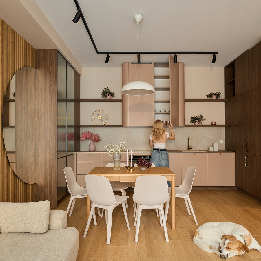 Aneks dzienny z kuchnią w pudrowych szafkach, drewniane akcenty, stół dla sześciu, lampa wisząca, sofa, śpiący pies, gość przy górnej szafce.