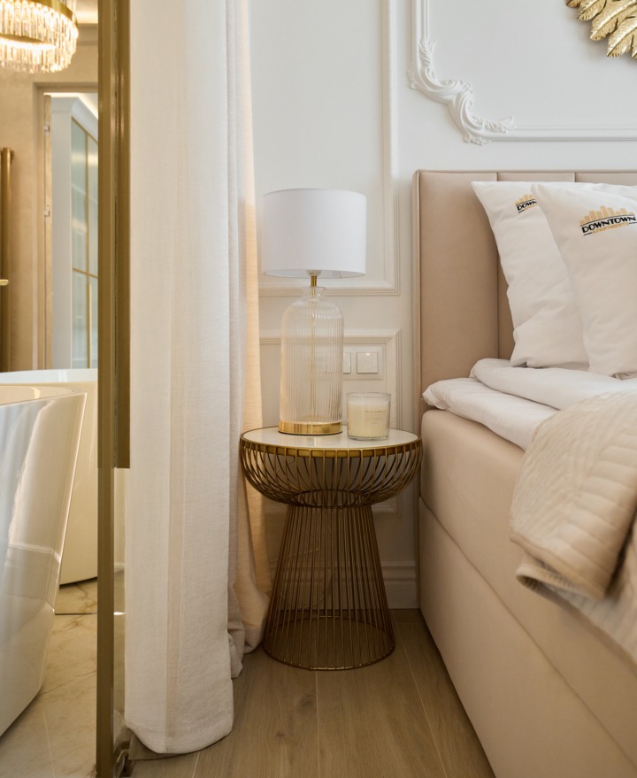 Beige Luxus-Hotellecke mit gepolstertem Bett, Kissen mit Logo, goldenem Draht-Beistelltisch, Glaslampe und freistehender Badewanne.