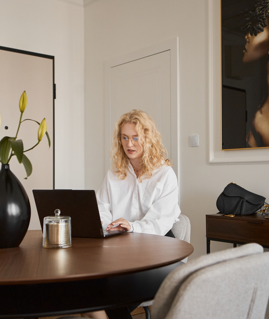 Gast arbeitet am Laptop an rundem Holztisch in eleganter Suite, Lilien in schwarzer Vase, moderne Wandkunst und Konsole mit Handtasche