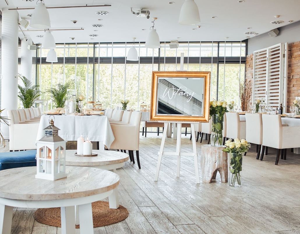 Bright event dining room with long tables covered in white tablecloths, cream chairs, wooden floors, large windows, welcome sign and flowers.