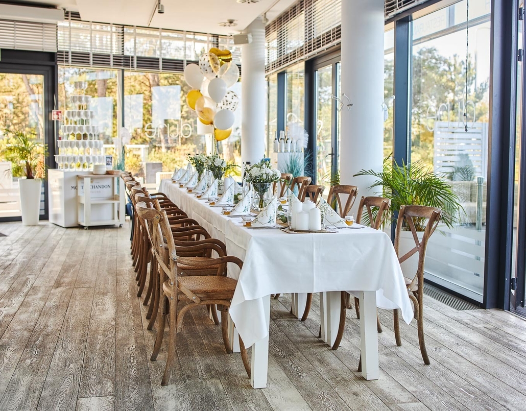 Bright, airy dining room set for a celebration: long white‑clothed table with wooden chairs, floral centerpieces, candles and balloons.