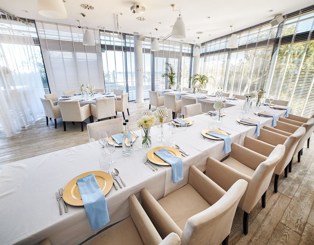 Sunlit modern banquet room with long tables draped in white linens, gold chargers, blue napkins and plush beige armchairs by floor-to-ceiling windows