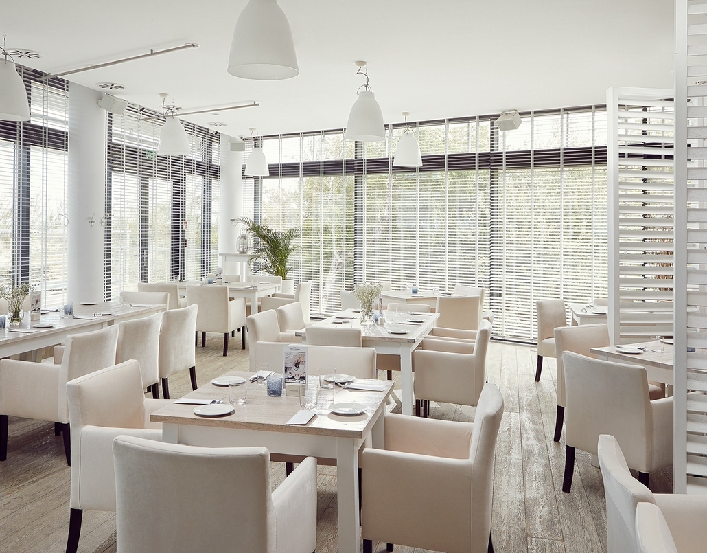 Bright modern hotel restaurant with cream upholstered chairs, wooden tables, pendant lights and floor-to-ceiling blinds