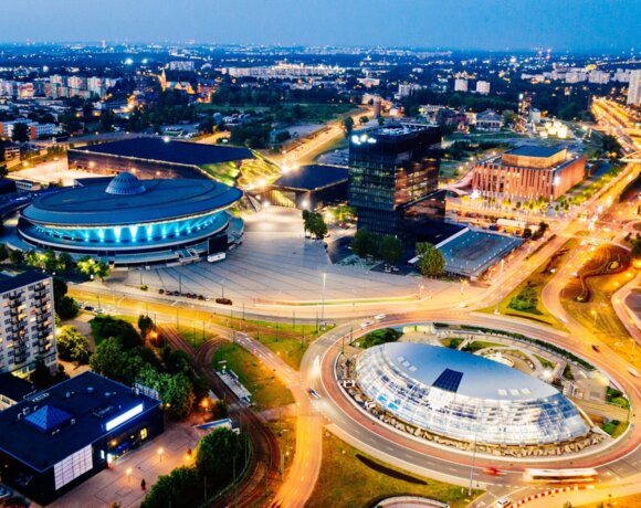 Night view of a modern city center with illuminated buildings, a roundabout, green areas, and urban infrastructure.