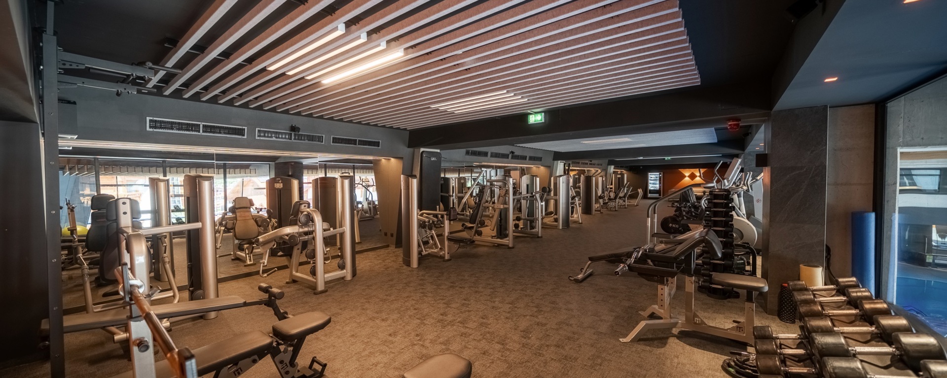 Modernes Hotel-Fitnessstudio mit Kraftgeräten, Hantelregal und Bänken, Spiegelwand, lamellenförmiger Holzdecke und warmem Licht