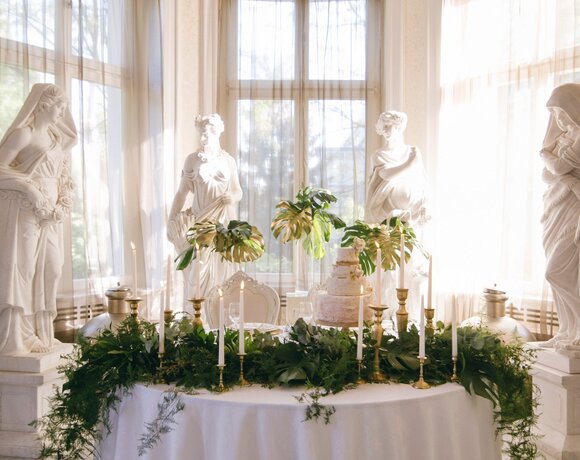 Sunlit reception table with three-tier wedding cake, brass candlesticks and greenery beneath a crystal chandelier and classical statues