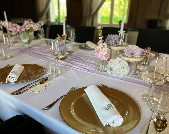 Elegancki stół bankietowy ze złotymi podtalerzami, monogramowanymi serwetkami, szkłem i pastelowymi kompozycjami kwiatowymi