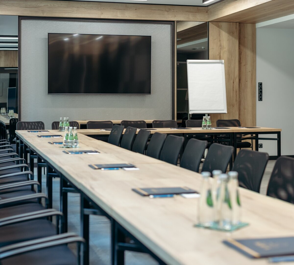 Moderner Hotel-Tagungsraum mit langen Holztischen, schwarzen ergonomischen Stühlen, großem Wand-TV, Flipchart und bereitgestellten Wasserflaschen.