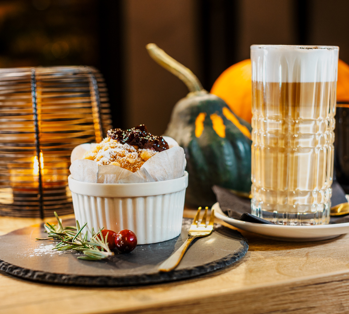 Herbstliches Dessert und Latte: Muffin mit Konfitüre und Puderzucker neben hohem Latteglas, Kerzenlicht und Kürbisdeko