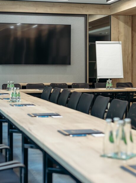Moderner Tagungsraum im Hotel mit langen Tischen, schwarzen Stühlen, wandmontiertem Bildschirm, Flipchart und Wasser.