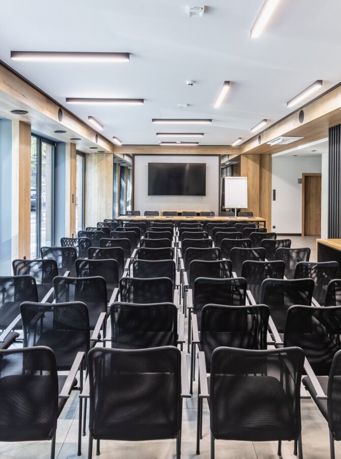 Moderner Hotel-Konferenzraum mit Reihen schwarzer Mesh-Stühle, großem Wanddisplay, bodentiefen Fenstern und warmen Holzakzenten