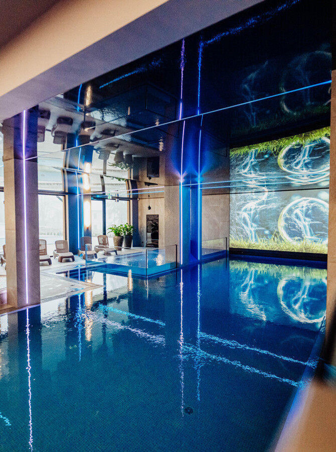 Innen-Spa im Hotel mit beleuchtetem Schwimmbecken, Whirlpool, Liegen, bodentiefen Fenstern und blauem LED-Ambiente