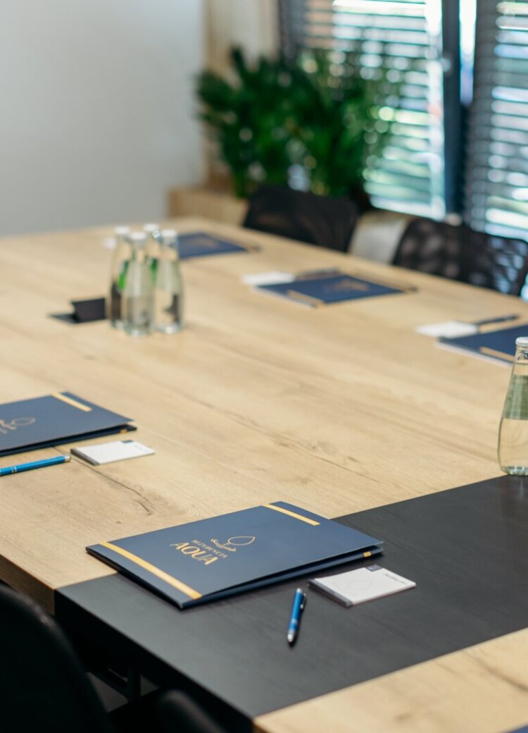 Konferenzraum mit langem Holztisch, vorbereitet für ein Meeting: Notizbücher, Stifte, Wasserflaschen und integrierte Steckdosen bei Jalousien.
