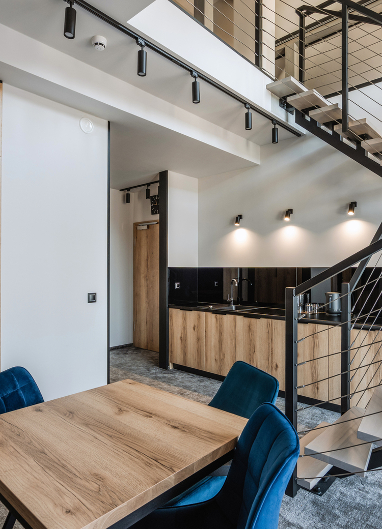 Modernes Duplex-Hotelzimmer mit Holz-Küche, Eßtisch und blauen Stühlen, metallene Treppe zur Galerie und sichtbarem Schlafzimmer.