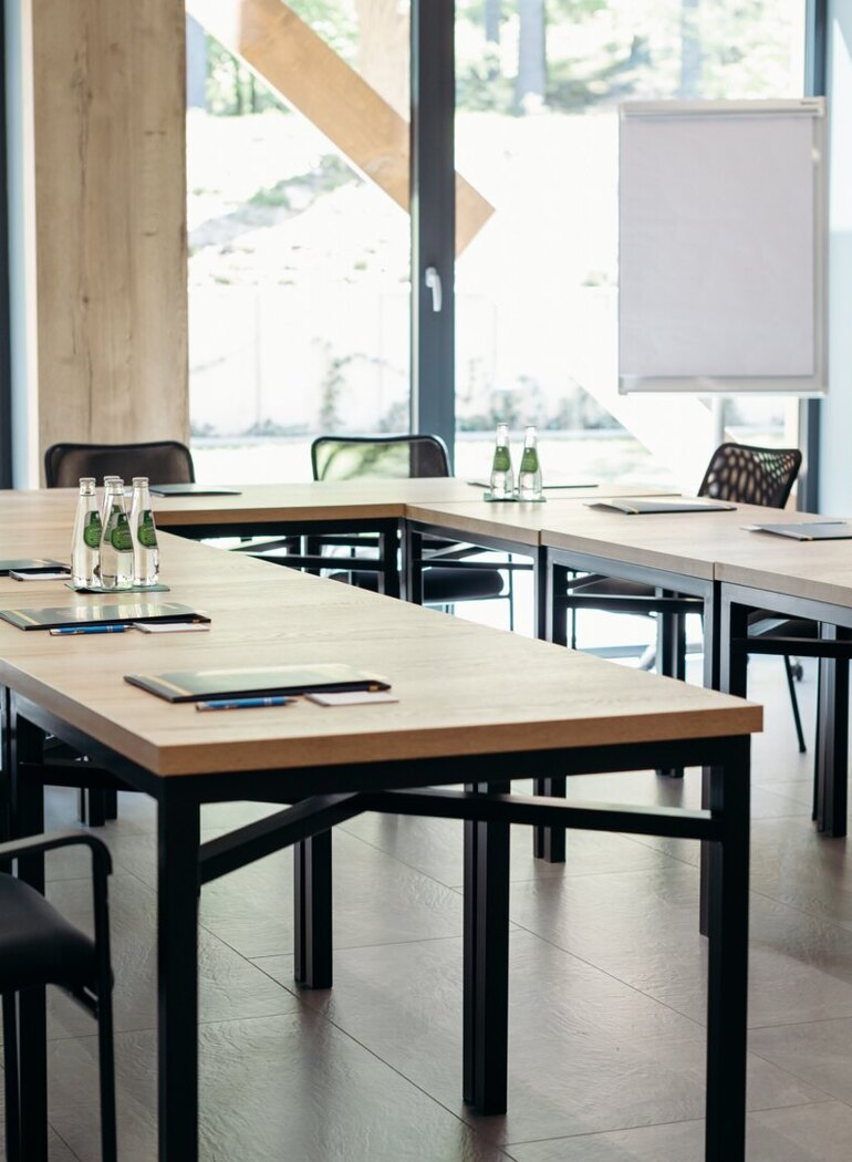 Konferenzraum in U‑Form mit Holztischen, bodentiefen Fenstern, Wasserflaschen, Notizblöcken, Stiften und Flipchart.