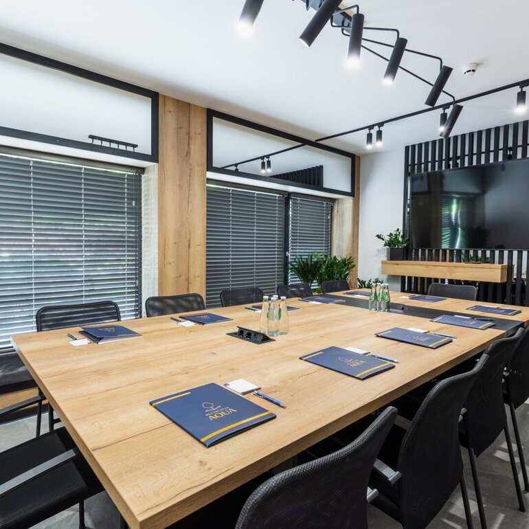 Moderner Hotel-Konferenzraum mit langem Holztisch, schwarzen Mesh-Stühlen, Marken-Notizblöcken, Wasserflaschen, Wand-TV, Jalousien und Pflanzen