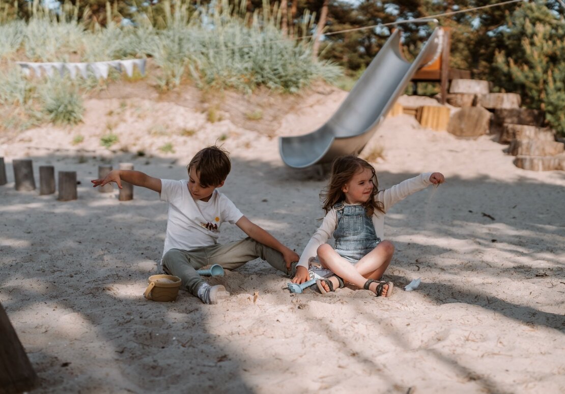 Two children playing in soft sand at a hotel's outdoor playground with a metal slide, dune grasses and bunting