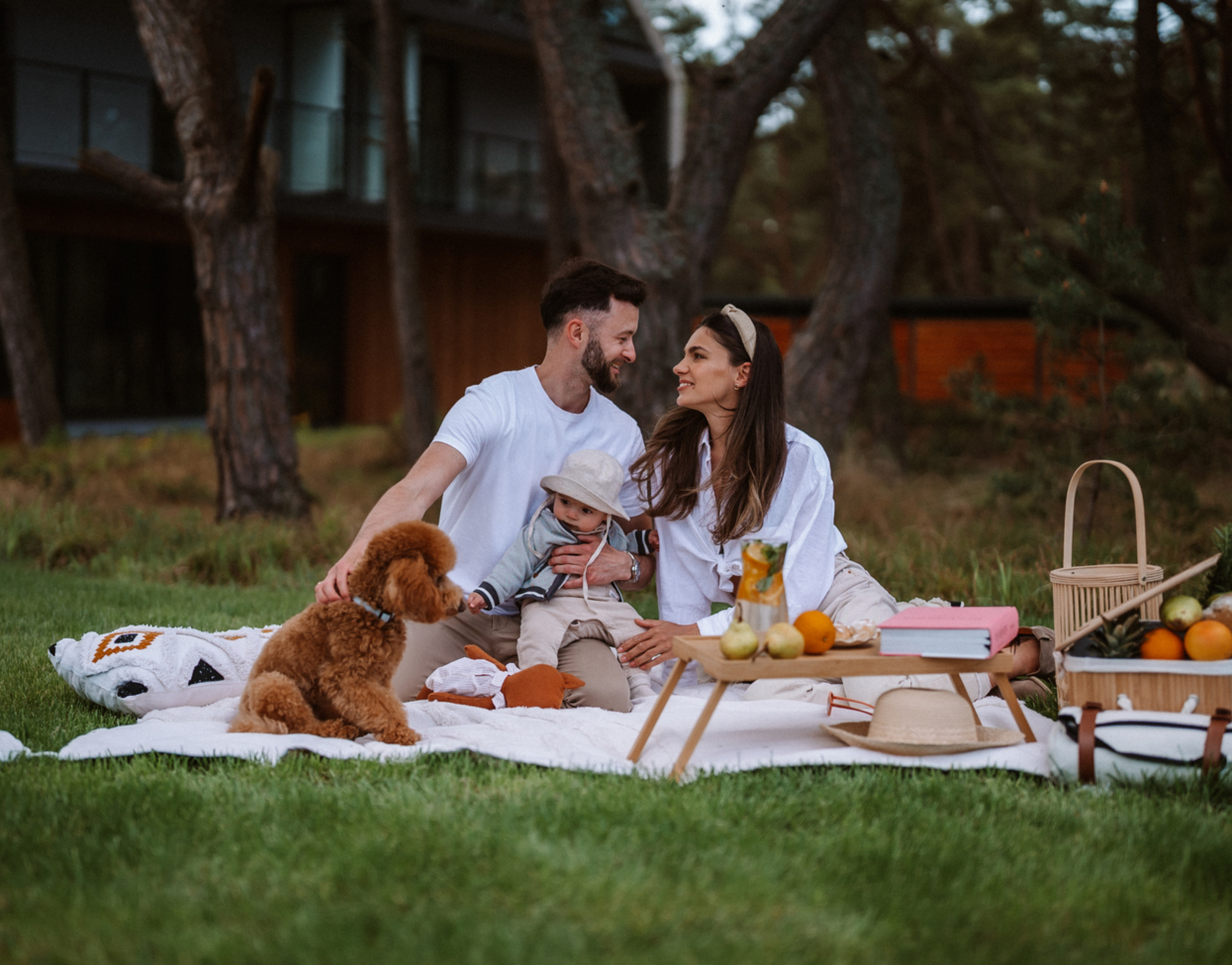 Family picnic on hotel lawn amid pine trees — couple with baby and dog on blanket, picnic basket, fruit and jug, modern lodge visible behind.
